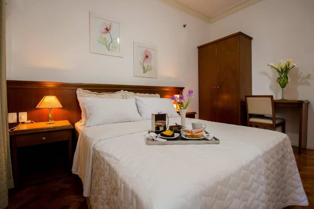 Uma cama bem arrumada com uma bandeja de café da manhã, criado-mudo, abajur, guarda-roupa, escrivaninha, cadeira e arte floral na parede em um quarto de hotel.