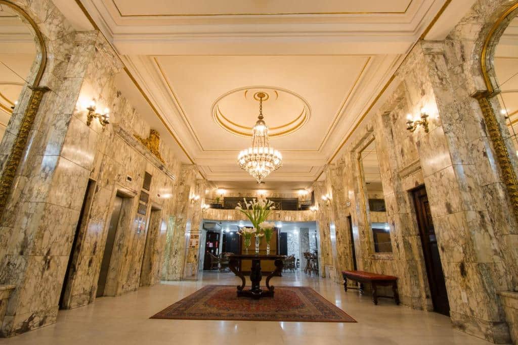 Grande lobby com paredes de mármore, lustres, uma mesa redonda central com flores e painéis espelhados nas paredes. Um dos hotéis no centro do rio janeiro.