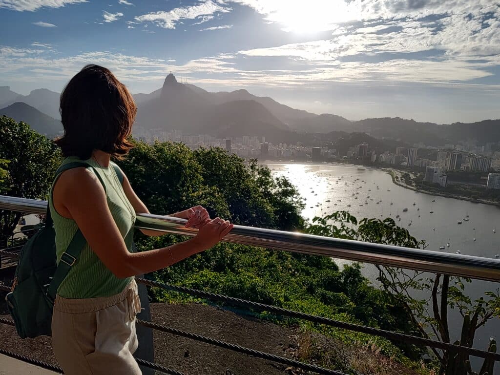 Uma pessoa está de pé perto de uma grade, com vista para uma paisagem urbana pitoresca com uma baía e montanhas sob um céu parcialmente nublado.