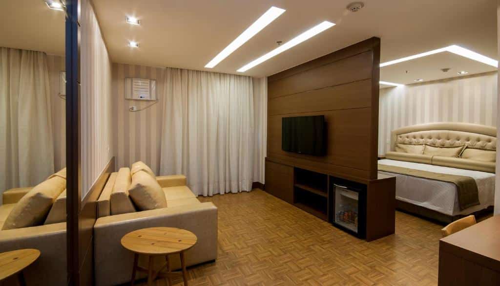 Quarto de hotel com um sofá bege, mesas redondas de madeira, uma TV em uma divisória e uma cama atrás da divisória. Este é um dos hotéis no centro do rio de janeiro.