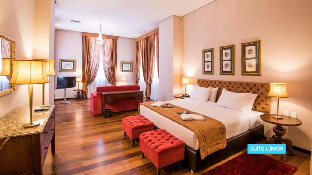 Quarto de hotel espaçoso com uma cama de casal, pufes vermelhos, um sofá, uma escrivaninha, luminárias, arte emoldurada e piso de madeira. Este é um dos hotéis no centro do rio de janeiro.