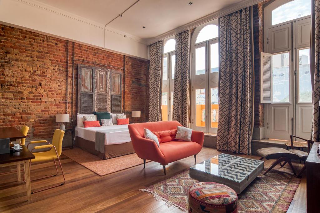 Um quarto espaçoso com uma cama, sofá laranja, tapete estampado, parede de tijolos, janelas grandes e piso de madeira. Este é um dos hotéis no centro do rio de janeiro.