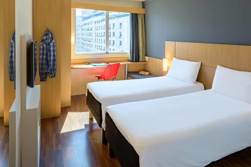 Quarto de hotel com duas camas de solteiro, piso de madeira, uma escrivaninha perto da janela, uma cadeira vermelha e camisas penduradas na parede.