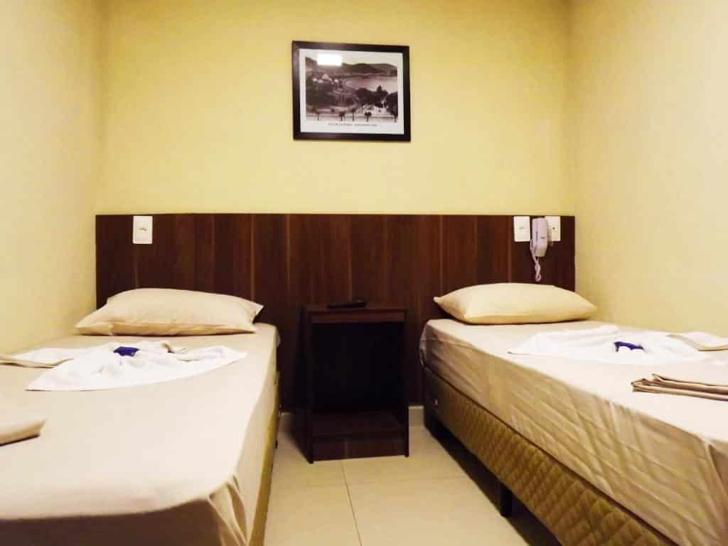 Duas camas de solteiro em um quarto pequeno com paredes bege, uma foto emoldurada e uma mesa de cabeceira entre as camas. Este é um dos hotéis no centro do rio de janeiro.