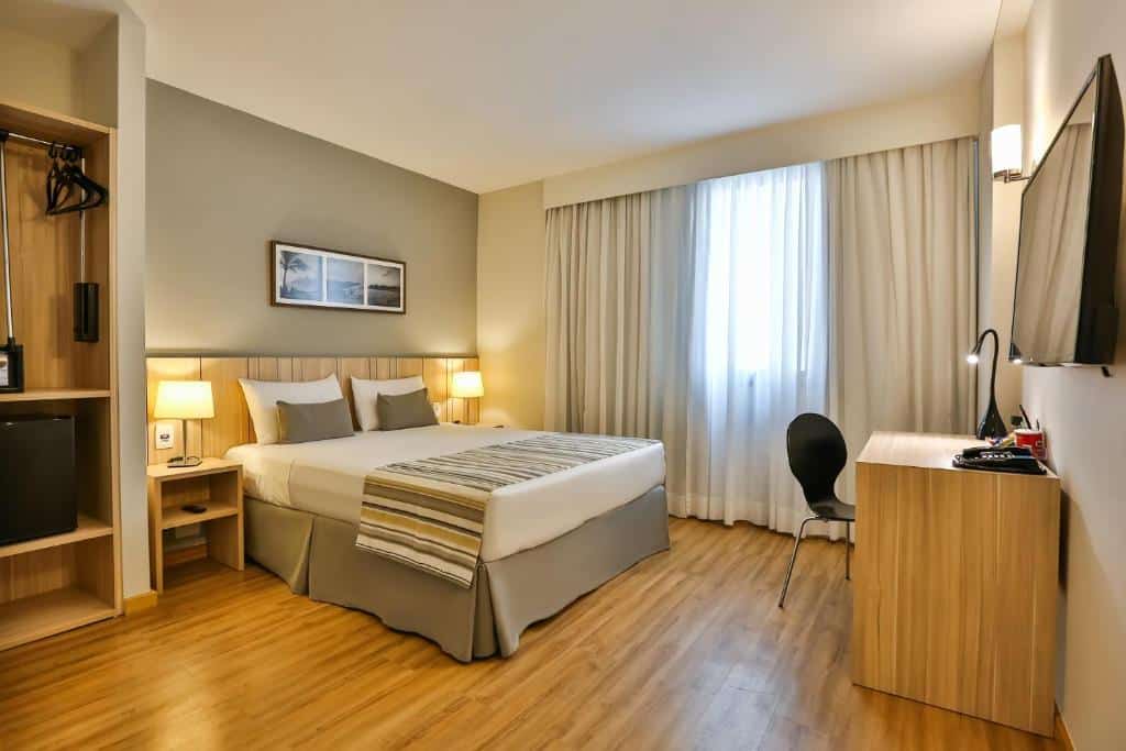 Quarto de hotel com uma cama de casal, duas mesinhas de cabeceira, escrivaninha, cadeira, TV, armário aberto e uma janela grande com cortinas.