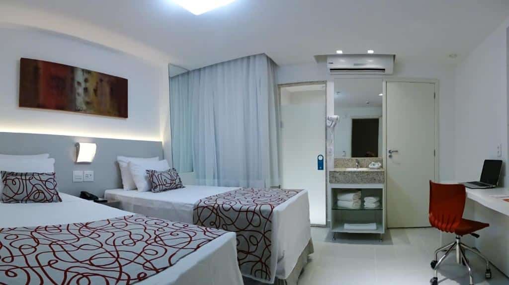 Quarto de hotel com duas camas de solteiro, uma escrivaninha com cadeira, área de pia e decoração branca com detalhes em vermelho. Este é um dos hotéis no centro do rio de janeiro.