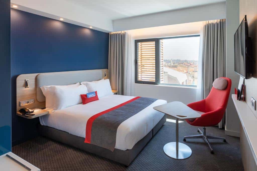 Quarto moderno no Holiday Inn Express Porto City Centre, an IHG Hotel com uma cama de casal, cadeira vermelha, mesa pequena, TV na parede e janela grande com vista para a cidade, para representar hotéis no Porto em Portugal.