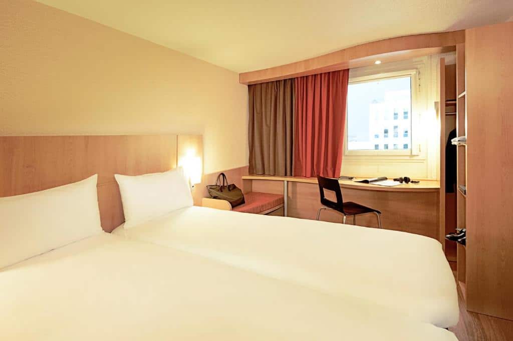 Quarto no ibis Porto Centro São Bento com duas camas de solteiro, uma janela, uma escrivaninha com uma cadeira, uma bolsa em um banco e móveis de madeira, para representar hotéis no Porto em Portugal.