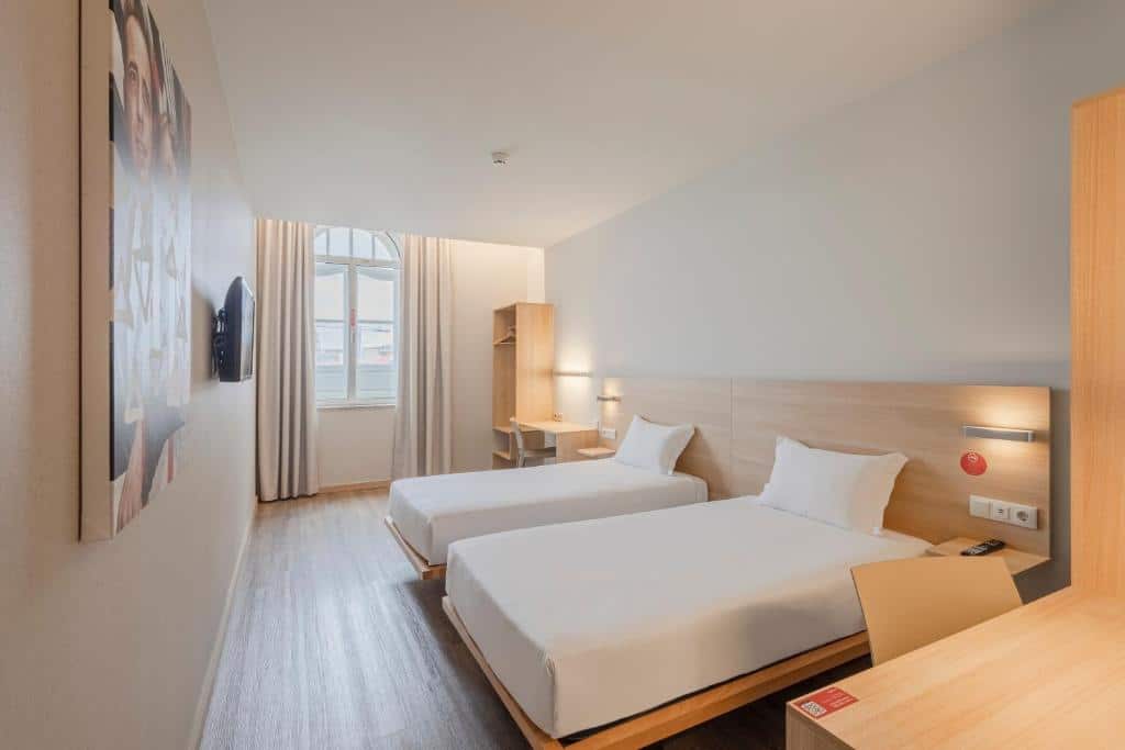 Quarto no Moov Hotel Porto Centro com duas camas de solteiro, uma escrivaninha, uma TV na parede e uma grande janela com cortinas que permitem a entrada de luz natural, para representar hotéis no Porto em Portugal.