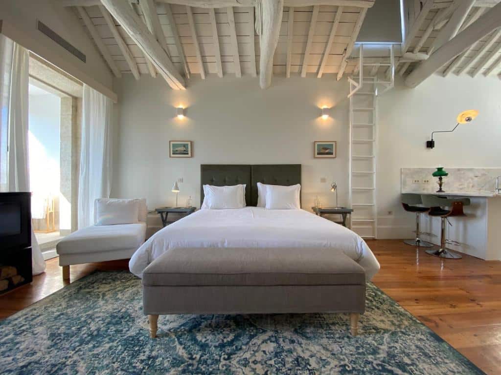 Um quarto moderno na Casa do Cativo com uma cama de casal, roupa de cama branca, uma espreguiçadeira, tapete e vigas expostas sob um teto alto, para representar hotéis no Porto em Portugal.