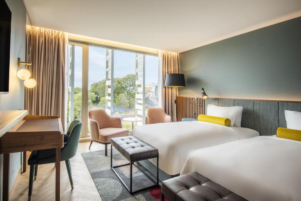 Quarto moderno no Renaissance Porto Lapa Hotel com duas camas, uma escrivaninha, cadeiras, janela grande e luz natural iluminando o espaço, para representar hotéis no Porto em Portugal.