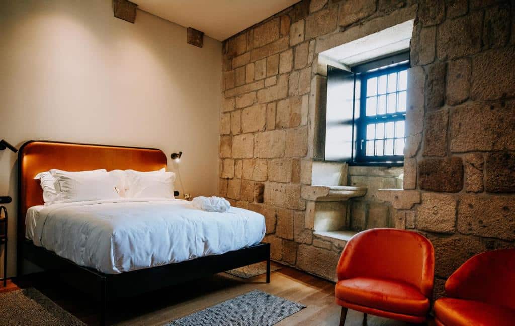 Um quarto moderno no Exmo. Hotel com uma cama de casal, duas cadeiras laranja, paredes de pedra e uma grande janela com grades pretas, para representar hotéis no Porto em Portugal.
