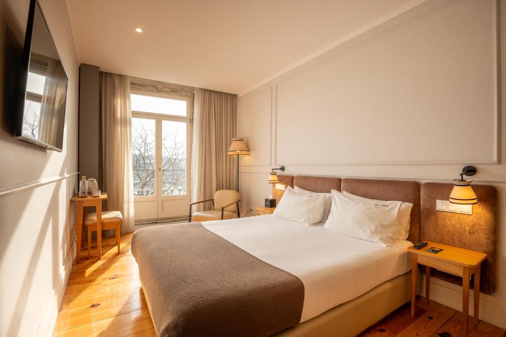 Um quarto do Memoria Porto FLH Hotels com uma cama de casal bem arrumada, duas mesinhas de cabeceira, uma escrivaninha, uma cadeira e uma grande janela que deixa entrar a luz do sol, para representar hotéis no Porto em Portugal.