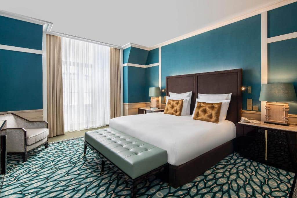 Um quarto moderno no Maison Albar Hotels Le Monumental Palace com uma cama de casal, paredes azul-petróleo, carpete estampado, poltrona, luminárias e uma grande janela com cortinas, para representar hotéis no Porto em Portugal.