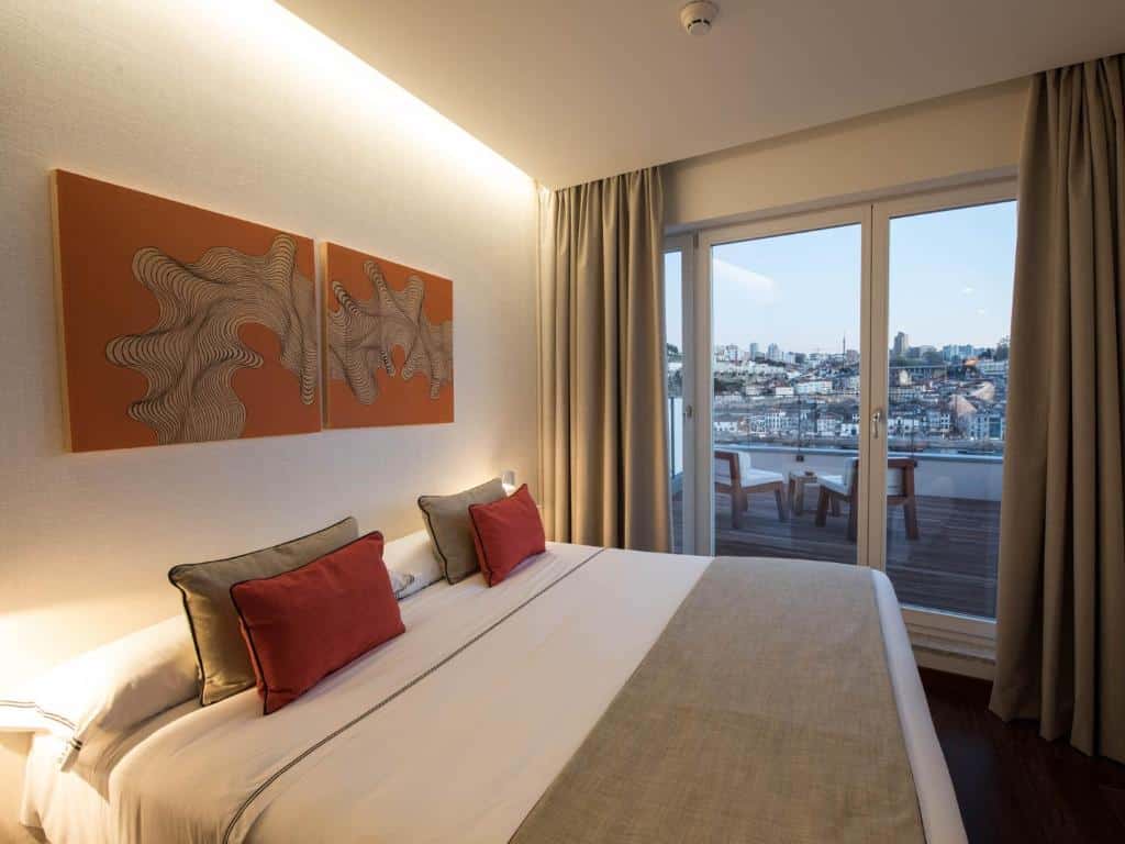 Quarto no Hotel Carris Porto Ribeira com uma cama grande, obras de arte em laranja, portas de vidro deslizantes e vista da paisagem urbana e da água na varanda, para representar hotéis no Porto em Portugal.