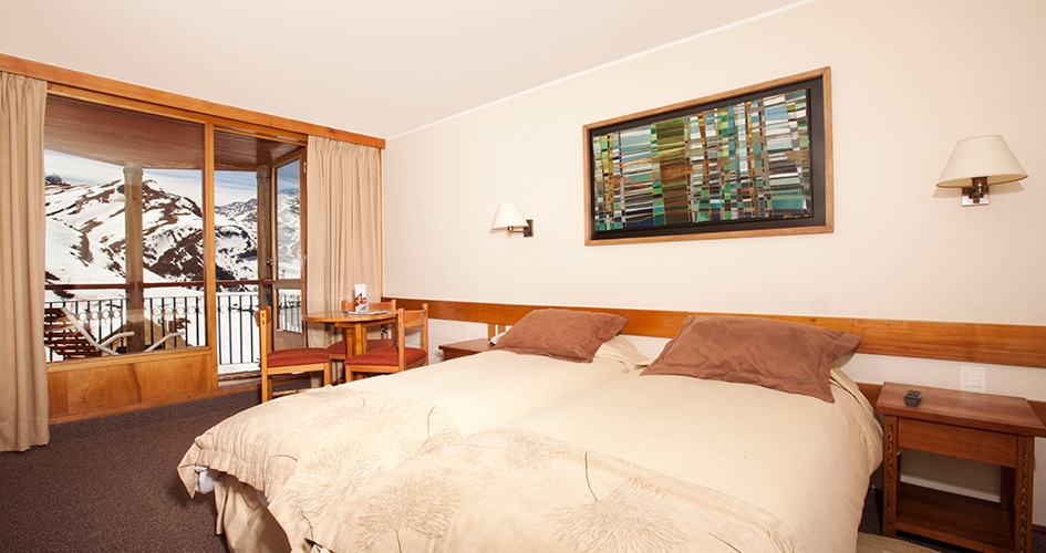 Quarto do Hotel Puerta del Sol com cama de casal do lado direito com duas cômodas ao lado da cama. Representa hotéis no Valle Nevado.