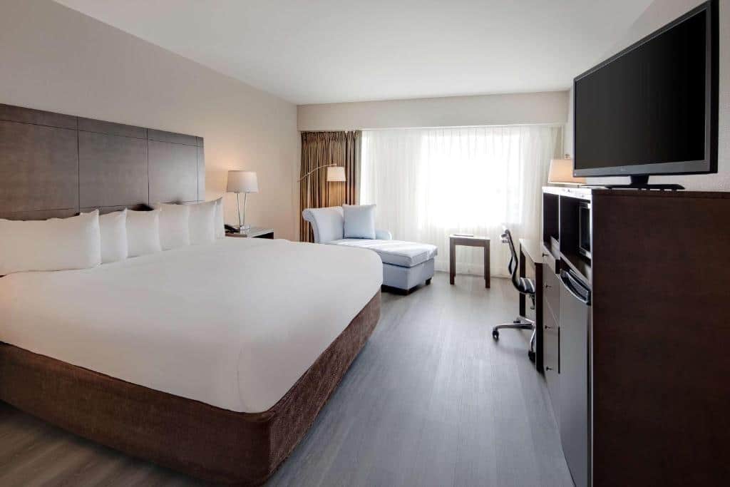 quarto do The Florida Hotel & Conference Center in the Florida Mall com cama de casal com luminárias de ambos os lados e tv na frente, com mesa e cadeiras mais ao fundo em frente a uma janela bem grande e um sofá em que é possível deitar