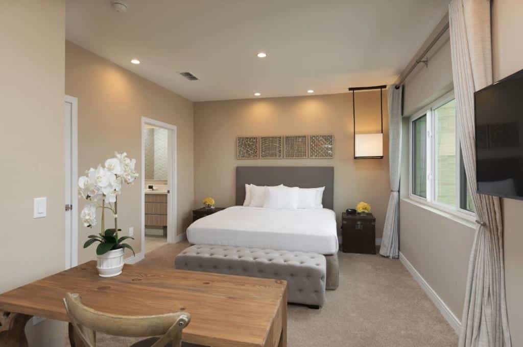 quarto de uma das villas do Magic Village Views Trademark Collection by Wyndham com mesinha, cama de casal com mesinha e luminária de ambos os lados, janela com cortinas e entrada para o banheiro em tons claros e refinados