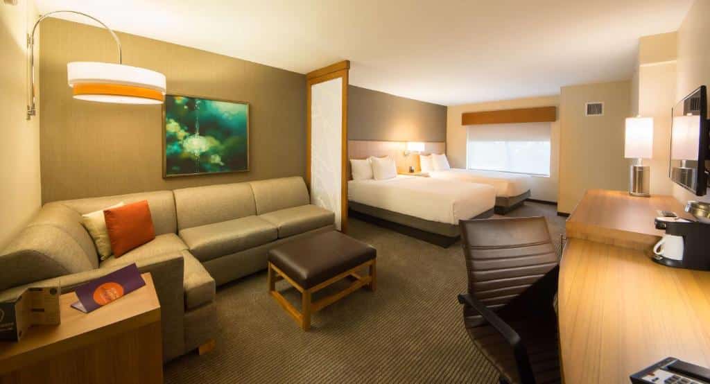 quarto grande com dois ambientes do Hyatt Place Orlando/Lake Buena Vista, um deles é uma sala de estar com sofá, mesa e tv e, no outro, cama de casal com janela grande e veneziana