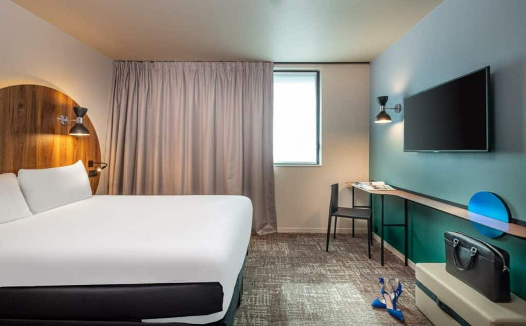 Quarto do ibis Styles Paris Meteor Avenue d'Italie, um dos hotéis para brasileiros em Paris. Uma cama de casal está ao lado esquerdo da imagem com mesinhas de cabeceira e luminárias dos dois lados. Na lateral direita da cama ficam duas janelas com cortinas, e logo em frente à cama há uma mesa comprida com cadeira, abajur e TV na parede acima.