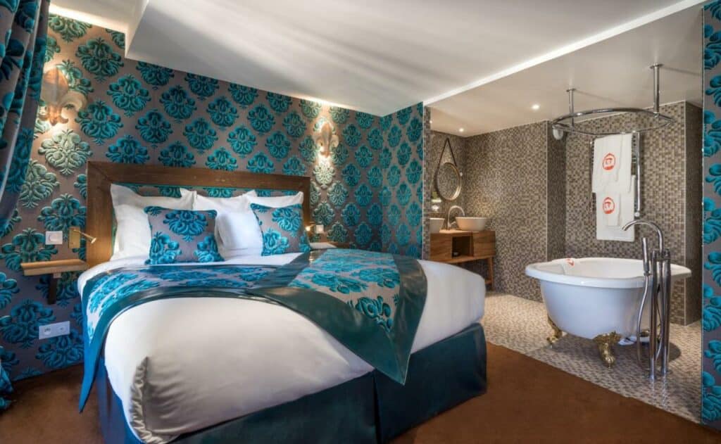 Quarto do Le Temple De Jeanne, um dos hotéis para brasileiros em Paris. Uma cama de casal está ao lado esquerdo do quarto e tem mesinhas de cabeceira dos dois lados. Na lateral direita fica o banheiro, onde é possível ver uma banheira de frente para as pias com espelhos.