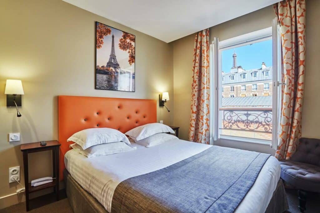 Quarto do Hôtel Le Relais Saint Charles, um dos hotéis para brasileiros em Paris. Uma cama de casal está ao lado esquerdo da imagem com mesinhas de cabeceira e luminárias dos dois lados. Na lateral direita da cama fica uma janela com cortinas e vista para a ponta da Torre Eiffel. Há um quadro do monumento na parede acima da cama.