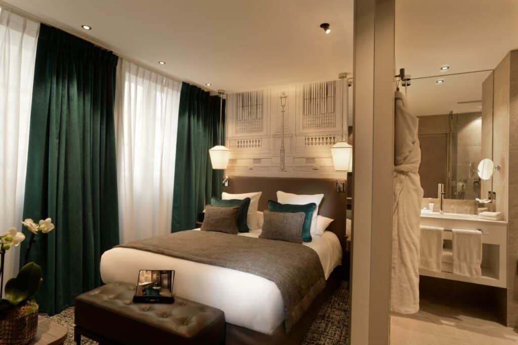 Quarto do Hotel La Lanterne & Spa, um dos hotéis para brasileiros em Paris. Uma cama de casal está ao lado esquerdo da imagem com mesinhas de cabeceira e luminárias dos dois lados. Na lateral esquerda da cama ficam duas janelas com cortinas. Uma parede no centro da imagem divide o quarto e mostra a pia do banheiro n=ao lado direito.