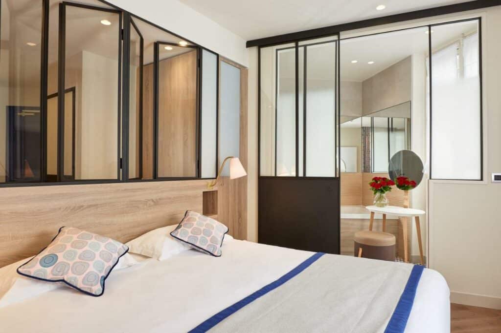 Quarto do Hôtel de Sévigné, um dos hotéis para brasileiros em Paris. Uma cama de casal está ao lado esquerdo da imagem com mesinhas de cabeceira e luminárias dos dois lados. Na lateral direita da cama fica uma porta que leva ao banheiro.