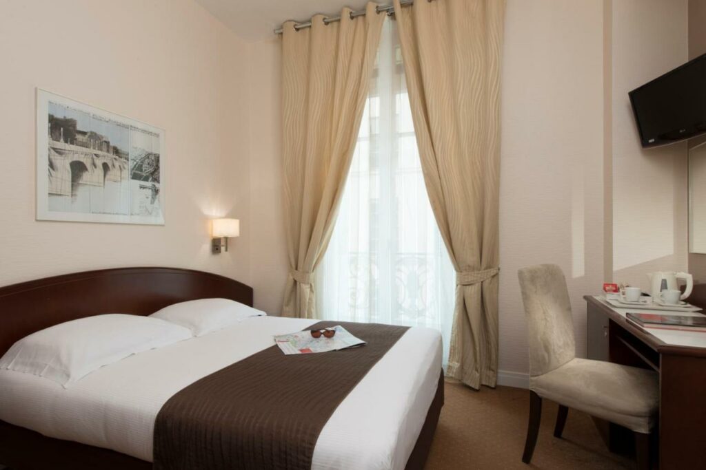 Quarto do Excelsior Opera, um dos hotéis para brasileiros em Paris. Uma cama de casal está encostada na parede ao lado esquerdo com mesinhas de cabeceira e luminárias dos dois lados, assim como um quadro logo acima. Ela encara uma TV na parede oposta, uma mesa com cadeira e comodidades para chá logo abaixo. A lateral direita da cama tem uma janela com cortinas.