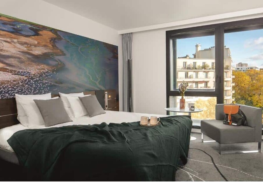 Quarto do Novotel Paris Porte Versailles, um dos hotéis para brasileiros em Paris. Uma cama de casal está encostada na parede do lado esquerdo e tem mesinhas de cabeceira e luminárias dos dois lados. Ao lado direito da imagem fica uma área de estar com mesinha e poltrona logo em frente à uma ampla janela com vista para a cidade.