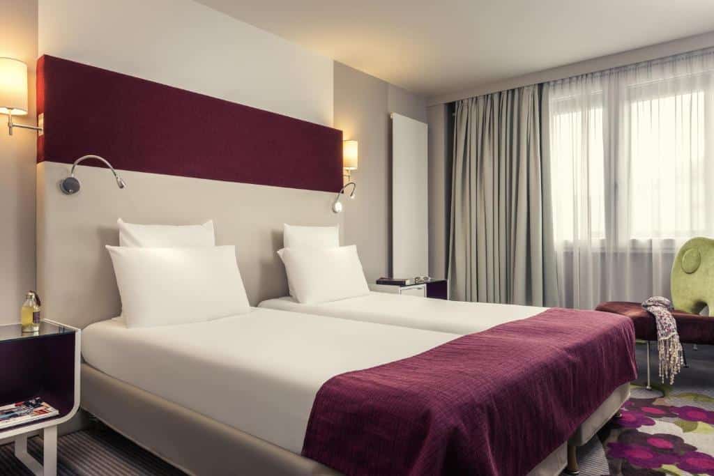 Quarto do Mercure Paris Bercy Bibliothèque, um dos hotéis para brasileiros em Paris. Uma cama de casal está ao lado esquerdo da imagem com mesinhas de cabeceira e luminárias dos dois lados. Na lateral direita da cama ficam duas janelas com cortinas, e logo em frente à elas há um banco acolchoado.