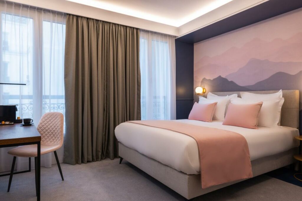 Quarto do Hôtel Le Milie Rose, um dos hotéis para brasileiros em Paris. Uma cama de casal está ao lado direito da imagem com mesinhas de cabeceira e luminárias dos dois lados. Na lateral esquerda da cama ficam duas janelas com cortinas, e logo em frente à cama há uma mesa de trabalho com cadeira e cafeteira.