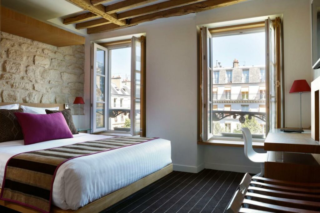 Quarto do Select Hotel, um dos hotéis para brasileiros em Paris. Uma cama de casal está ao lado esquerdo da imagem com mesinhas de cabeceira com luminárias dos dois lados. Na lateral direita da cama ficam duas janelas com cortinas, e logo em frente à cama há uma mesa de trabalho com cadeira, abajur e TV na parede acima.