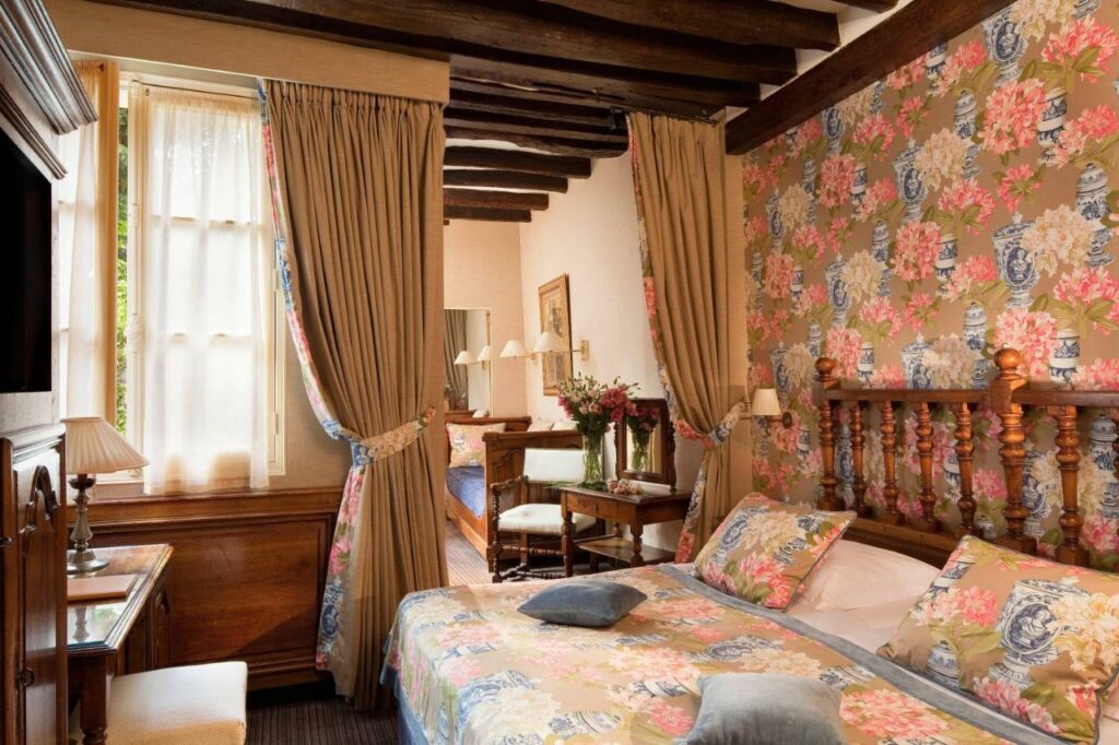 Quarto do Hotel Left Bank Saint Germain, um dos hotéis para brasileiros em Paris. Uma cama de casal está ao lado direito da imagem com luminárias dos dois lados. Em frente fica uma mesa de trabalho com cadeira, abajur e TV na parede acima. Na lateral esquerda da cama fica uma área de estar com poltrona e sofá. Os cômodos são separados por uma cortina.