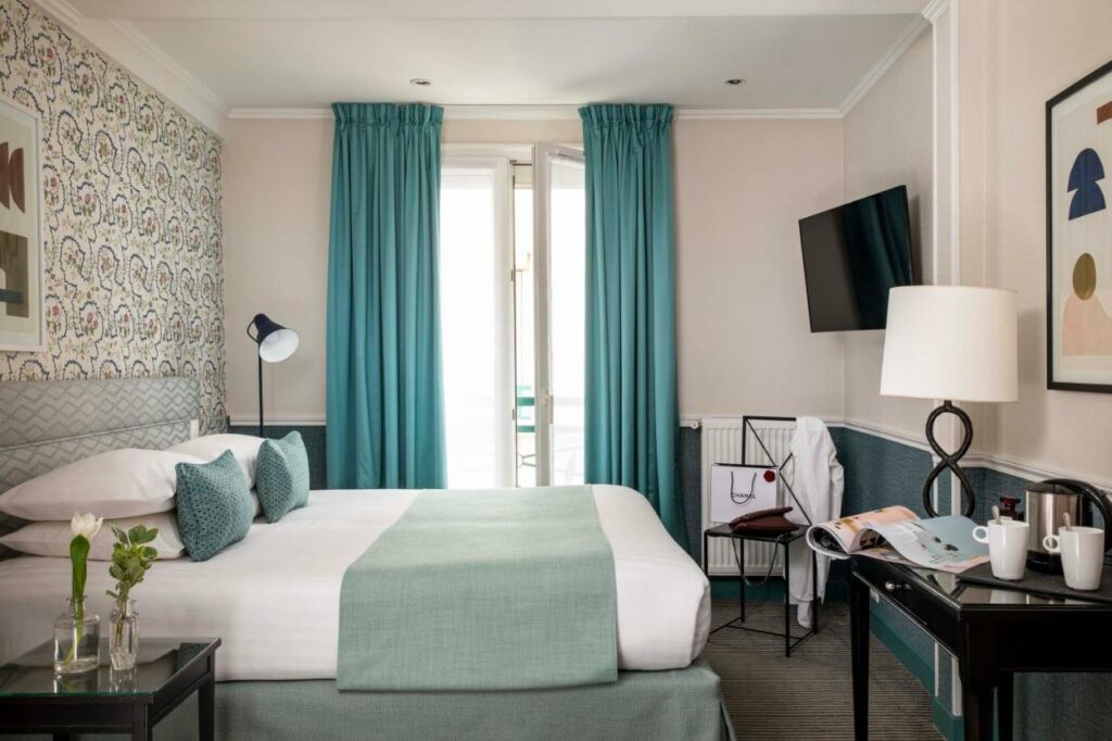 Quarto do Hotel Relais Bosquet by Malone, um dos hotéis para brasileiros em Paris. Uma cama de casal está ao lado esquerdo da imagem com mesinhas de cabeceira e luminárias dos dois lados. Na lateral direita da cama fica uma janela com cortinas, e logo em frente à cama há uma mesa com cafeteira, abajur e TV na parede acima.