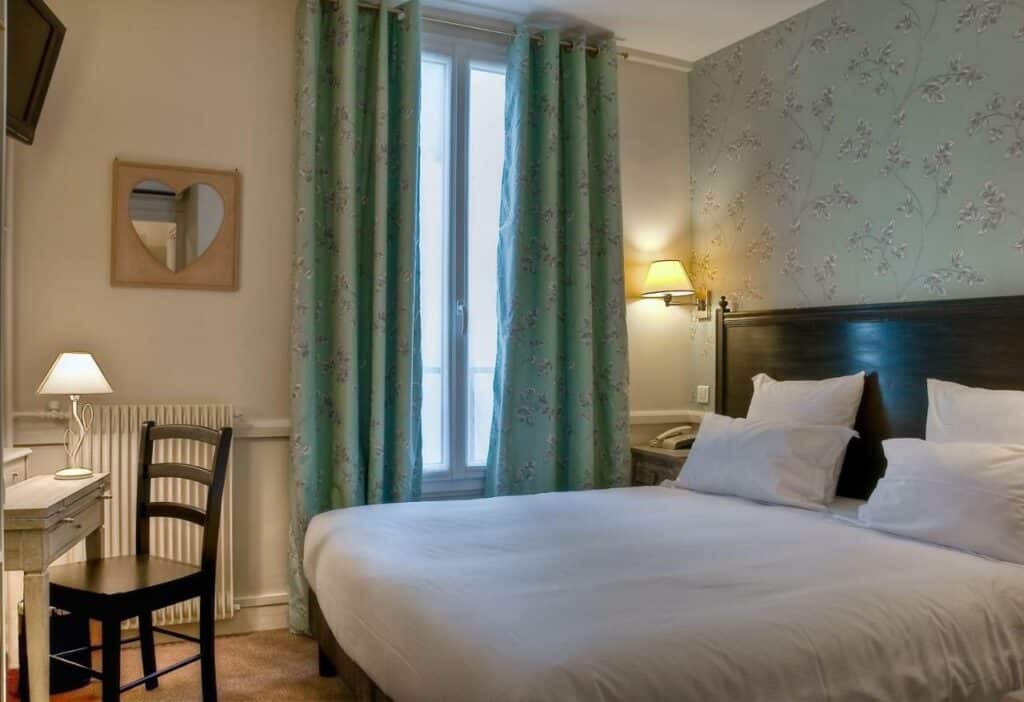 Quarto do Hôtel de la Motte Picquet, um dos hotéis para brasileiros em Paris. Uma cama de casal está ao lado direito da imagem com mesinhas de cabeceira e luminárias dos dois lados. Na lateral esquerda da cama fica uma janela com cortinas, e logo em frente à cama há uma mesa de trabalho com cadeira, abajur e TV na parede acima. Ao lado da janela há um espelho em formato de coração.