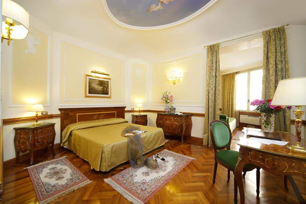 quarto do Hotel Hiberia, um dos hotéis bem localizados para brasileiros em Roma, com cama de casal, mesinha e luminária de ambos os lados, mesa e poltrona à frente, com tapetes no chão de madeira que combina com todos os móveis, também em madeira, há uma janela grande com cortinas e decoração clássica