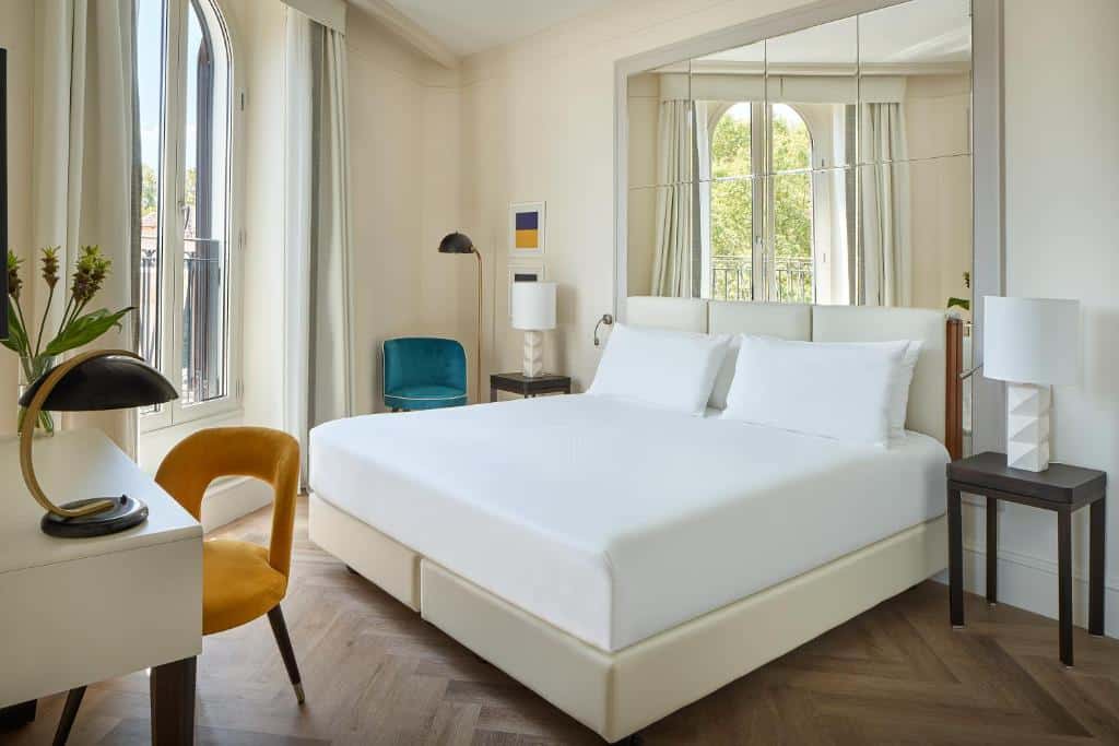 quarto do UNAHOTELS Trastevere Roma, um dos hotéis para brasileiros em Roma, com cama de casal, mesinha e luminária de ambos os lados, poltronas, mesa com cadeira, duas janelas grandes com cortinas claras