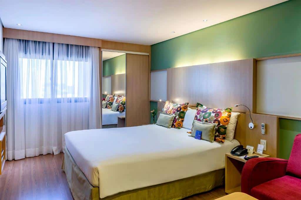 Quarto do Mercure Sao Paulo Naçoes Unidas com uma janela com cortinas, um pequeno armário de madeira com um espelho, uma cama de casal com almofadas e travesseiros, e do lado esquerdo, um móvel com uma televisão, o chão é de madeira, para representar hotéis para o The Town em SP