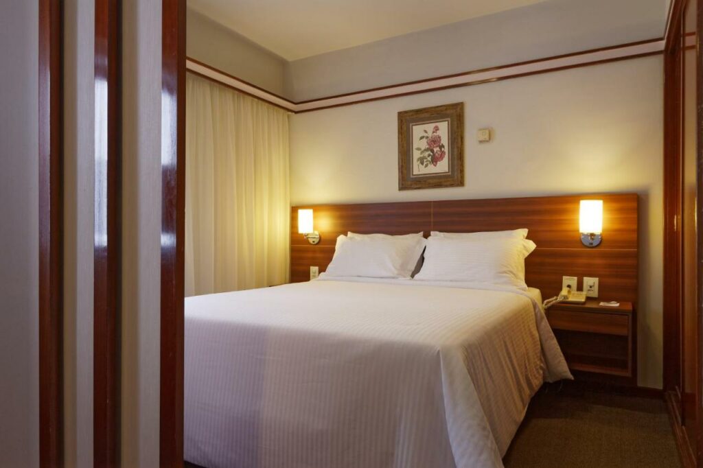 Quarto do Bristol The Time Berrini com uma cama de casal, uma janela do lado esquerdo com cortinas e um armário do lado direito