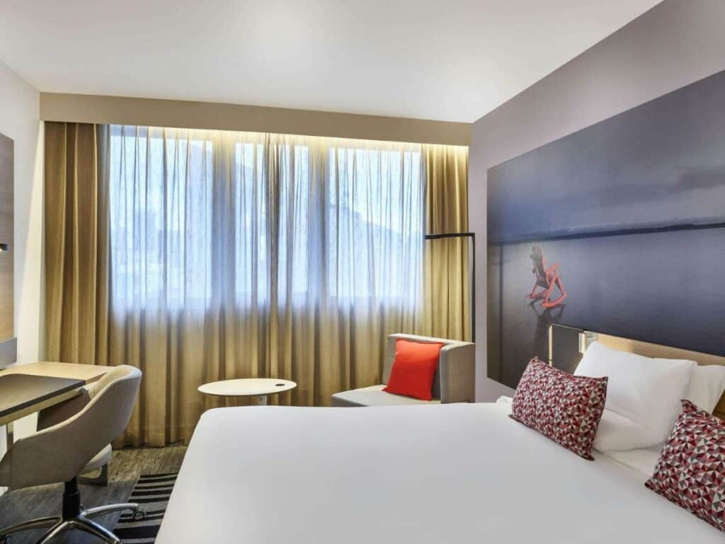 Quarto no Novotel Sao Paulo Morumbi com uma cama de casal, uma janela ampla com cortinas, com uma poltrona e uma mesinha do lado, e de frente com a cama há uma mesinha de escritório com uma cadeira giratória