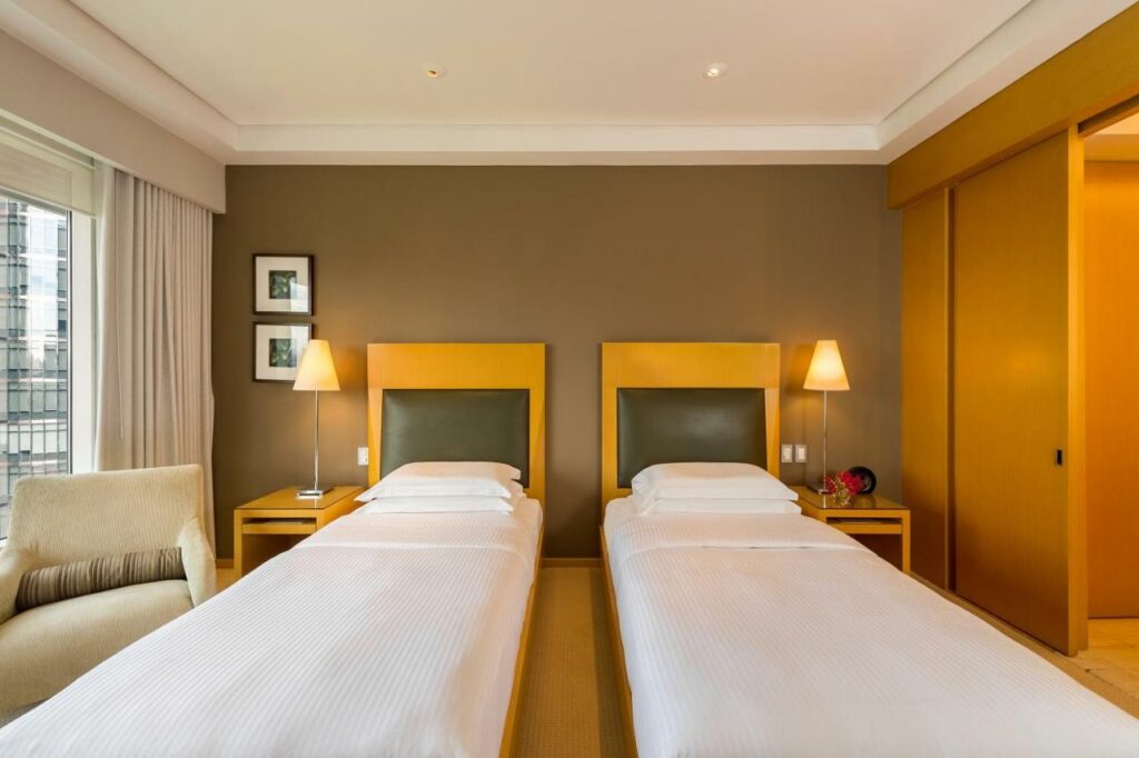 Quarto no Grand Hyatt São Paulo com duas camas de solteiro, do lado esquerdo tem uma janela com cortinas e uma poltrona estofada, o chão é de madeira e há duas mesinhas de madeira com abajures sob elas nas laterais das camas, para representar hotéis para o The Town em SP