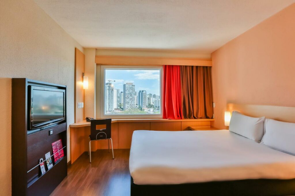 Quarto no ibis Sao Paulo Congonhas com uma janela com cortinas e embaixo dela há uma mesa de escritório com uma cadeira, do lado direito está uma cama de casal e de frente para ela tem uma televisão, o chão é de madeira