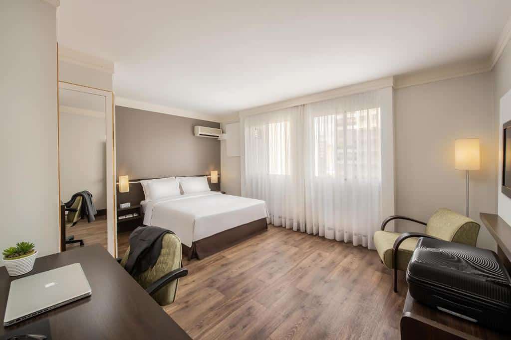 Quarto amplo no Intercity São Paulo Nações Unidas com uma janela grande com cortinas, uma cama de casal, um espelho de corpo inteiro, uma mesa de escritório com uma cadeira giratório, uma televisão, uma poltrona e um abajur de chão, o chão é de madeira, para representar hotéis para o The Town em SP