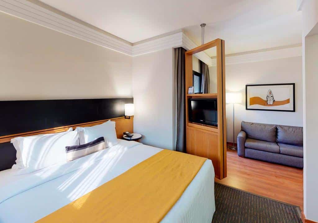 Quarto no Estanplaza International com uma cama de casal, do lado direito há uma televisão e um sofá com dois lugares, o chão é de madeira e há uma pequena luminária do lado da cama, para representar hotéis para o The Town em SP