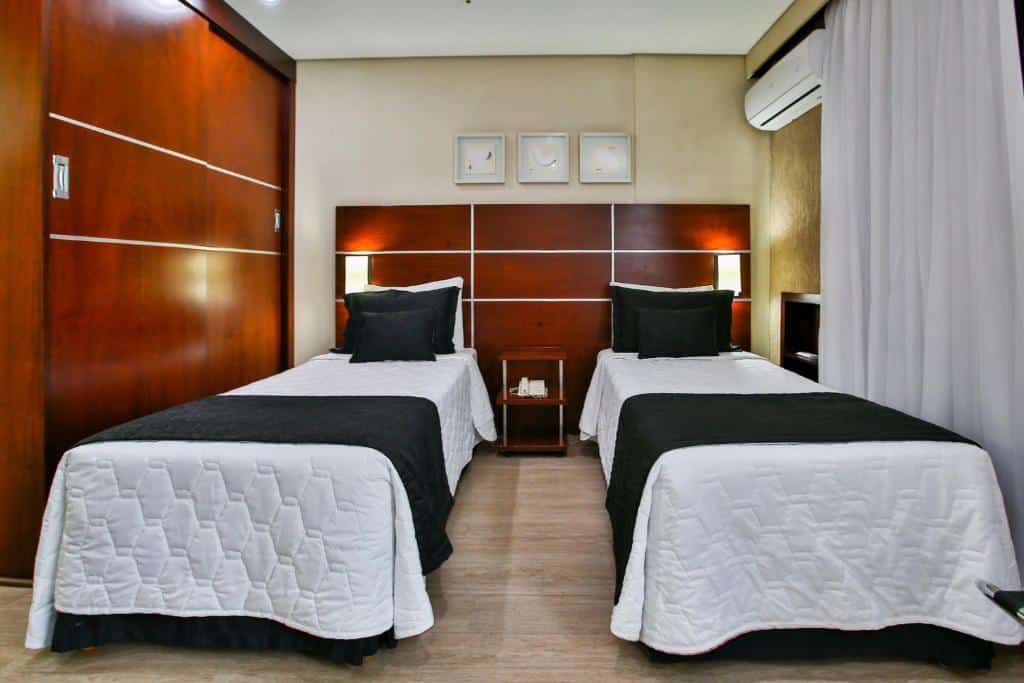 Quarto no Transamerica Executive Chácara Santo Antônio com duas camas de solteiro, do lado direito há uma janela com cortinas e um ar-condicionado, o chão é de madeira e há pequenas luminárias nas laterais das camas, para representar hotéis para o The Town em SP