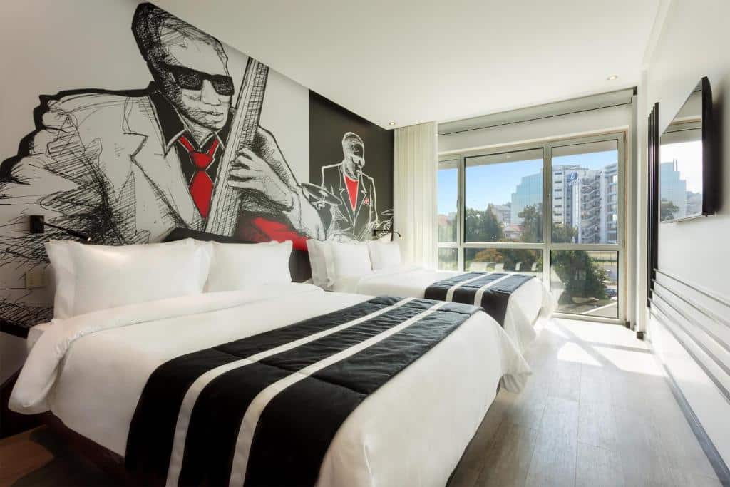 Quarto espaçosos do HF Fenix Music com duas camas de casal, uma varanda panorâmica com cortinas, e de frente para as camas há uma televisão, para representar hotéis perto da Praça Marquês de Pombal em Lisboa