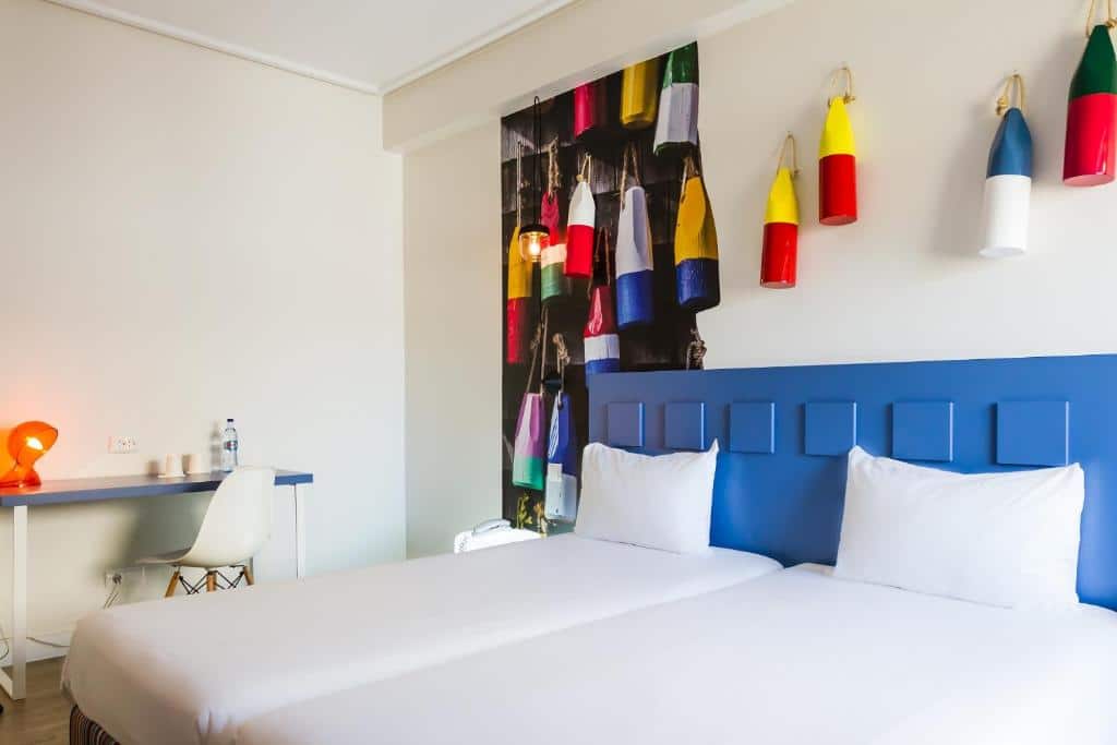 Quarto do Hotel Ibis Styles Lisboa Liberdade com duas camas de solteiro, uma pequena mesa com uma luminária e uma cadeira, há uma decoração em azul e com quadros coloridos sob as camas