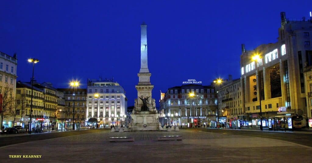 Praça dos Restauradores em Lisboa, uma praça muito espaçosa com prédios antigos ao redor e, no centro, há um monumento.