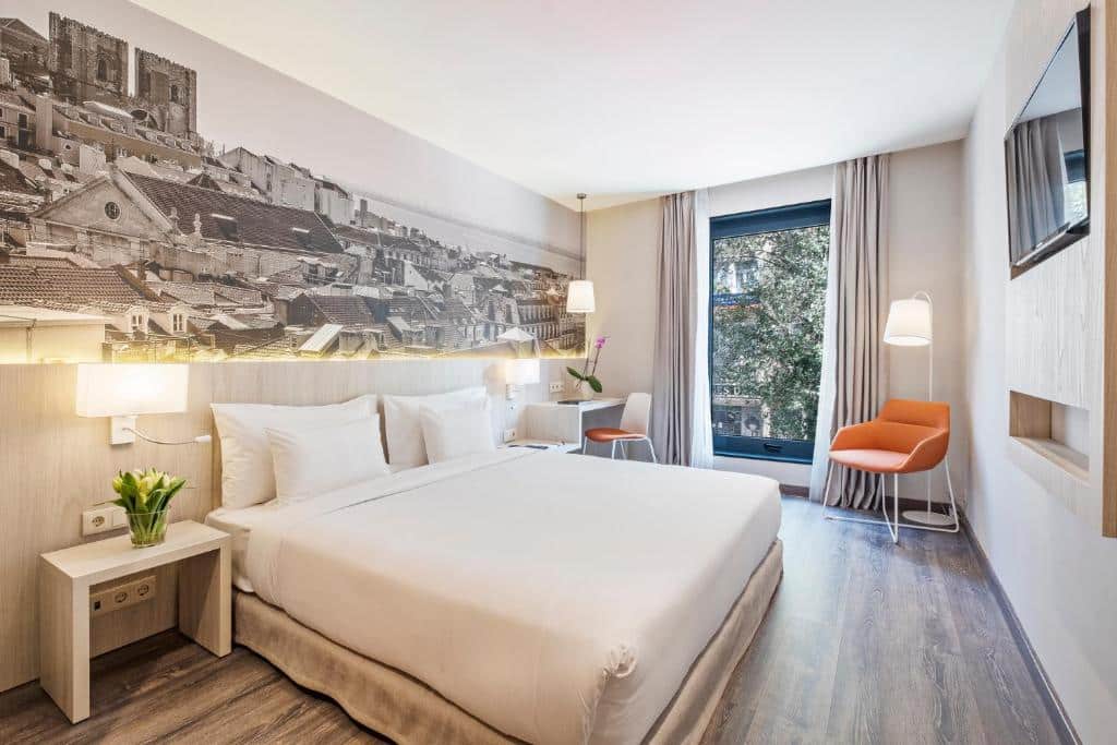 Quarto do EXE Liberdade com uma janela ampla com cortinas, próximo dela há uma poltrona com uma luminária de chão, assim como uma mesa de escritório com uma cadeira, há uma cama de casal e, de frente para ela, há uma televisão, para representar hotéis perto da Praça Marquês de Pombal em Lisboa